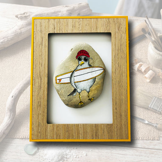 Galet peint à la main représentant une mouette avec un bonnet rouge et une planche de surf, cadre bois avec contour jaune, artisanat d'art L'Atelier du Ressac Auvergne.